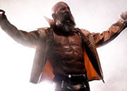 A muscular bearded male wrestler or performer stands with arms raised in a dramatic pose on a smoky stage, wearing a dark jacket with orange lining and black pants.
